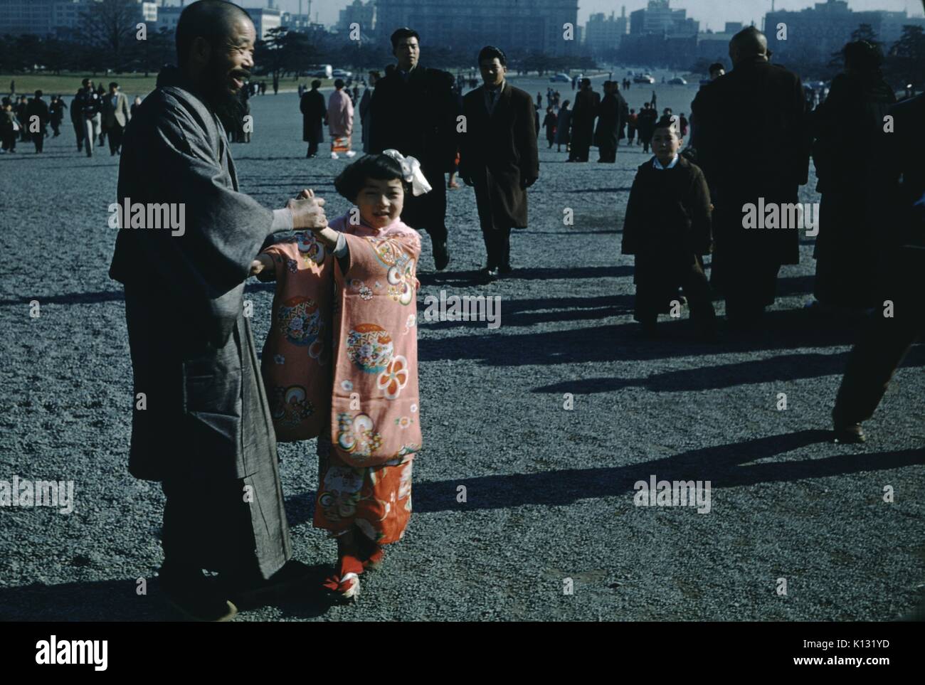 L'homme japonais avec tête à tête, et robe longue barbe de sourire et de tenir les mains de sa jeune fille, qui porte un kimono rose traditionnel et Sandales Geta, sur une place publique, avec un grand nombre de personnes à pied passé en groupes de deux à trois, 1952. Banque D'Images