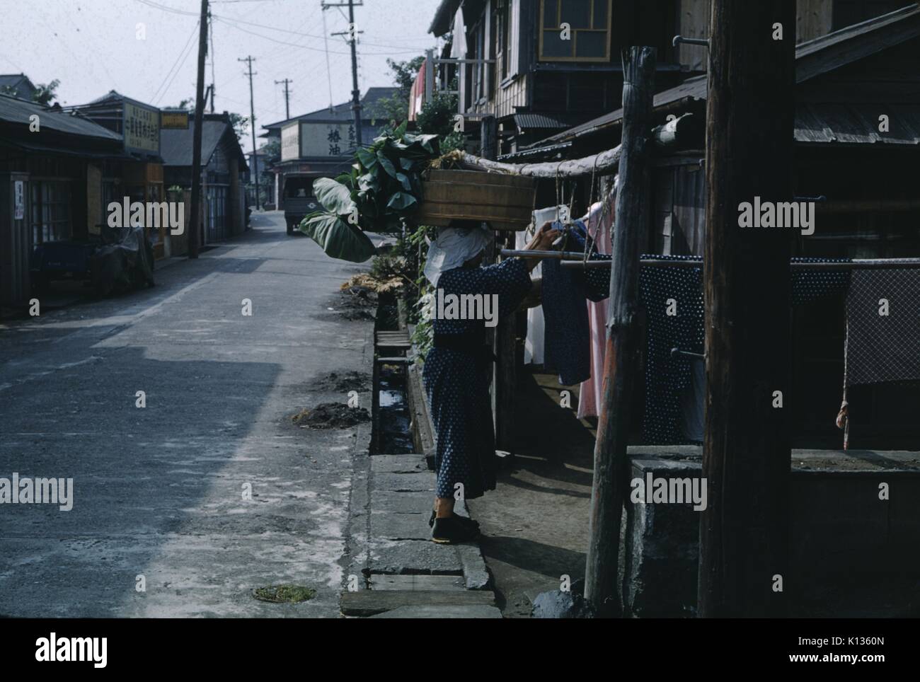 Femme portant un grand panier sur sa tête tandis qu'étendre le linge sur une corde à l'avant d'une maison sur une rue traditionnelle, l'île de Oshima, Japon, 1951. Banque D'Images