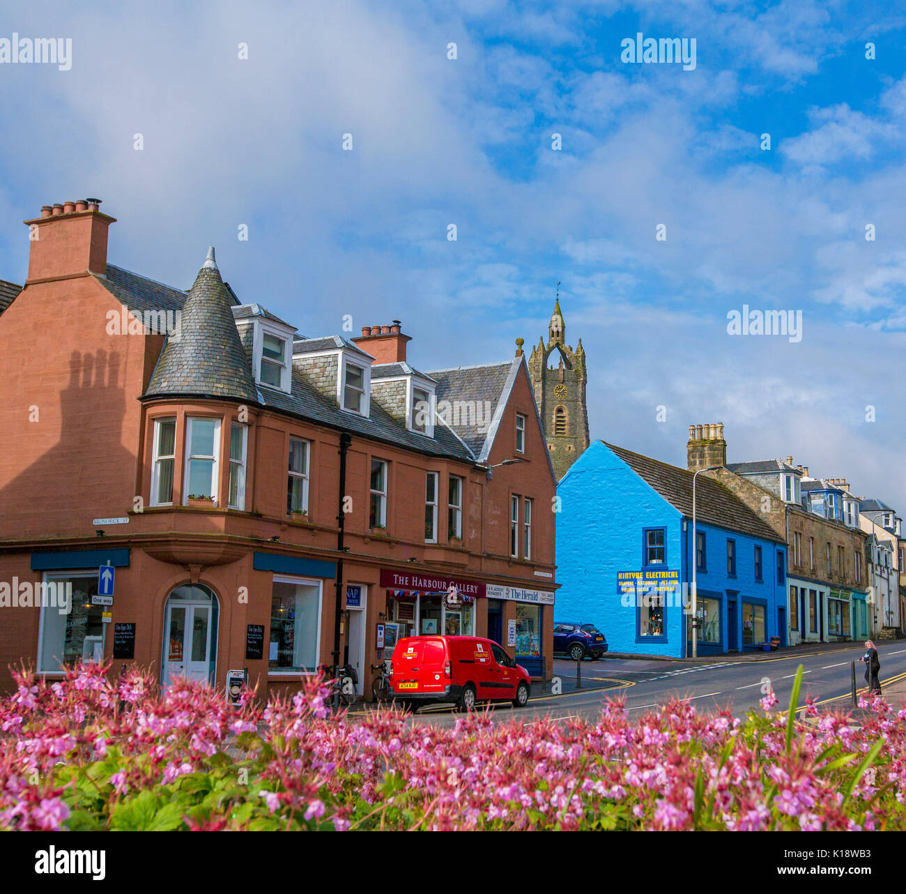 Village écossais de Tarbert avec rangée de bâtiments aux couleurs vives / boutiques bordant la rue principale et des fleurs roses en premier plan under blue sky Banque D'Images