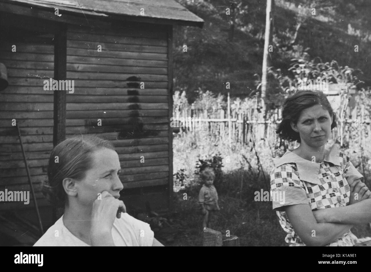 Deux femmes et un petit enfant, dans une ville minière, avec une simple maison, envahie par l'herbe, et une clôture en bois construite environ servant de toile de fond, la Virginie de l'Ouest, 1935. À partir de la Bibliothèque publique de New York. Banque D'Images