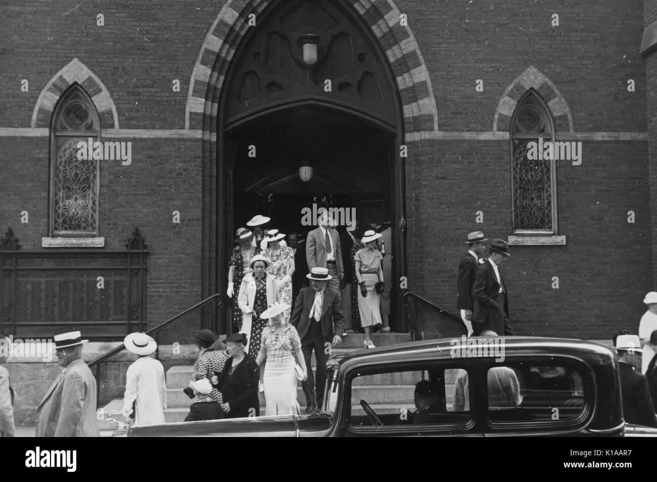 Groupe d'hommes bien habillés et les femmes, les hommes en costumes et chapeaux, les femmes en robes et chapeaux, gants, à quitter une église, voiture garée en face, Manchester, New Hampshire, 1937. À partir de la Bibliothèque publique de New York. Banque D'Images