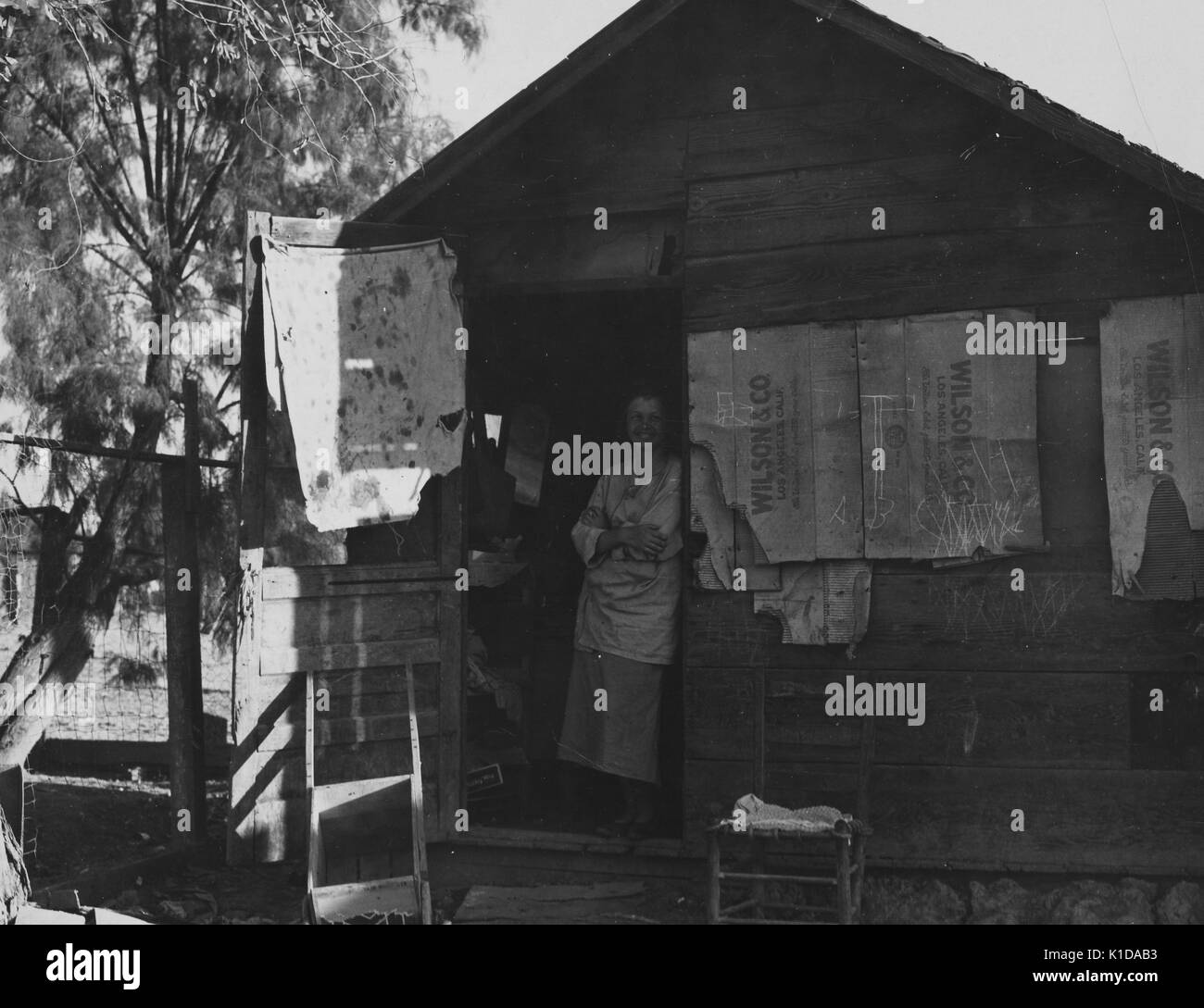 Femme immigrée souriante debout à l'entrée d'une cabane en bois, une chaise à l'extérieur, fenêtres couvertes de caisses de Wilson Company, Californie, 1935. De la bibliothèque publique de New York. Banque D'Images