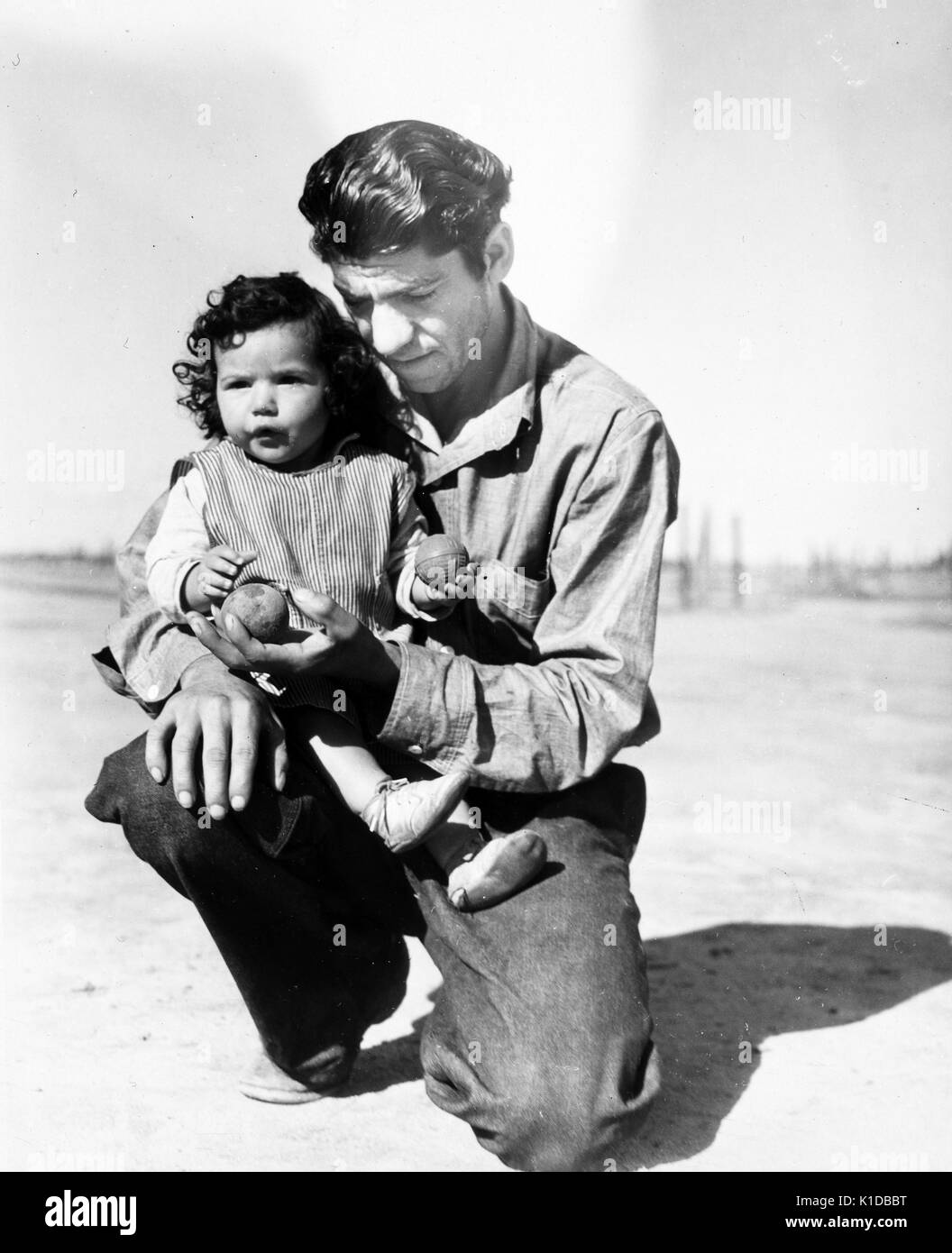 Travailleur migrant à genoux et tenant sa fille, regardant vers le bas avec une expression du visage grave, très éclairé avec des ombres, Californie, 1936. à partir de la bibliothèque publique de new york. Banque D'Images