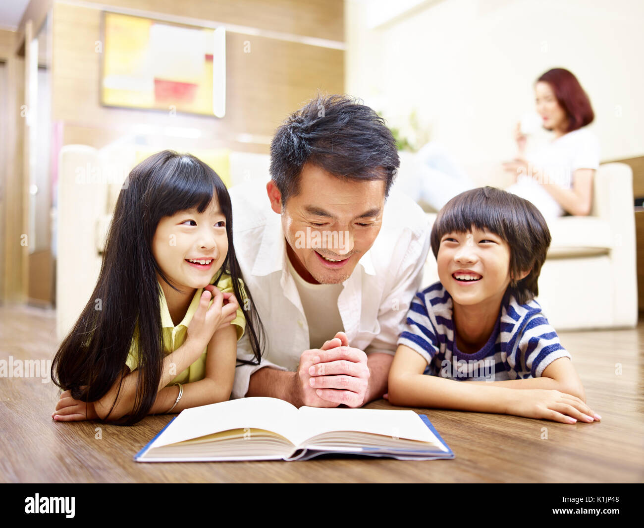 Le père et les enfants asiatiques lying on floor reading book avec la mère à l'arrière-plan. Banque D'Images