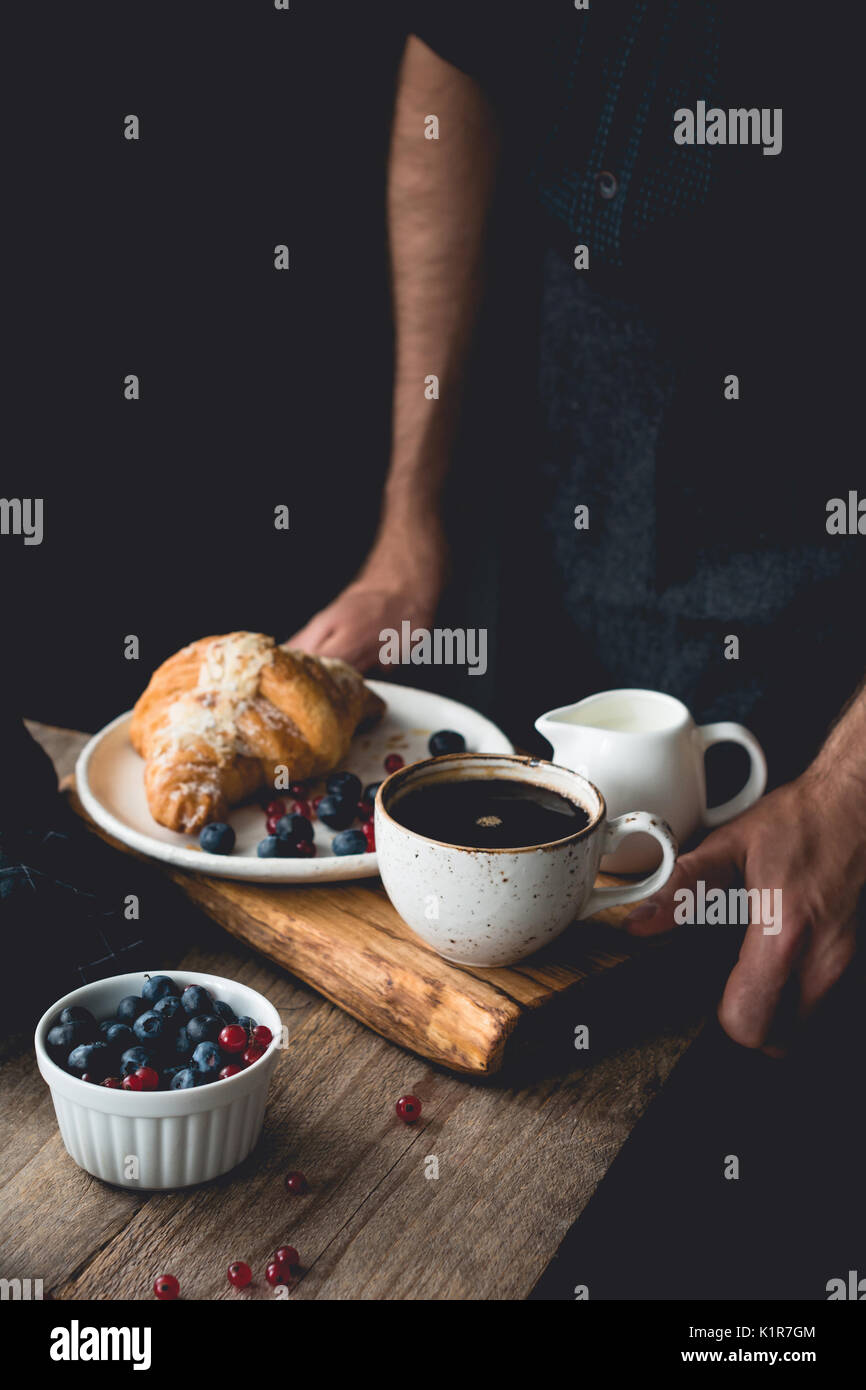 Petit déjeuner sur un plateau : croissants, café, crème et petits fruits. man hands holding plateau en bois avec petit déjeuner Banque D'Images