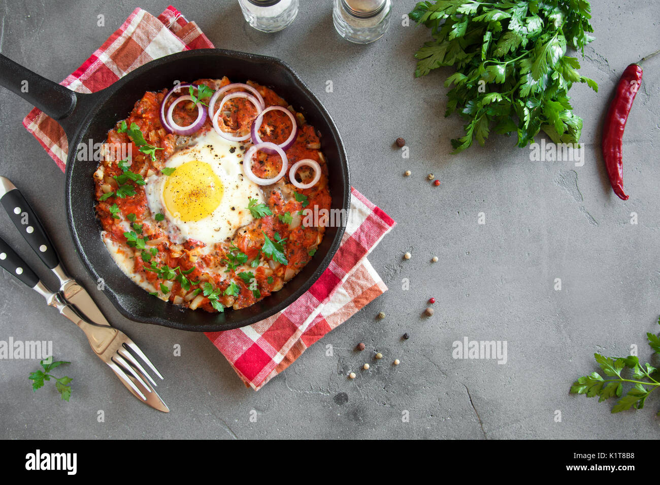 Shakshuka dans une poêle en fonte. Style moyen-orientale oeufs pochés dans une sauce tomate et du poivre avec le persil, l'oignon et les épices sur fond de béton gris. Banque D'Images