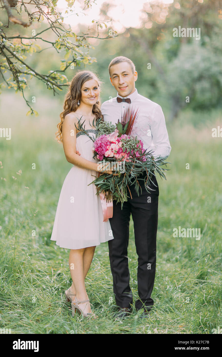 L'ensemble de l'entreprise portrait de la demoiselle souriante dans le genou-longueur robe tenue l'énorme bouquet et le meilleur homme souriant serrant avec elle au fond de la forêt. Banque D'Images