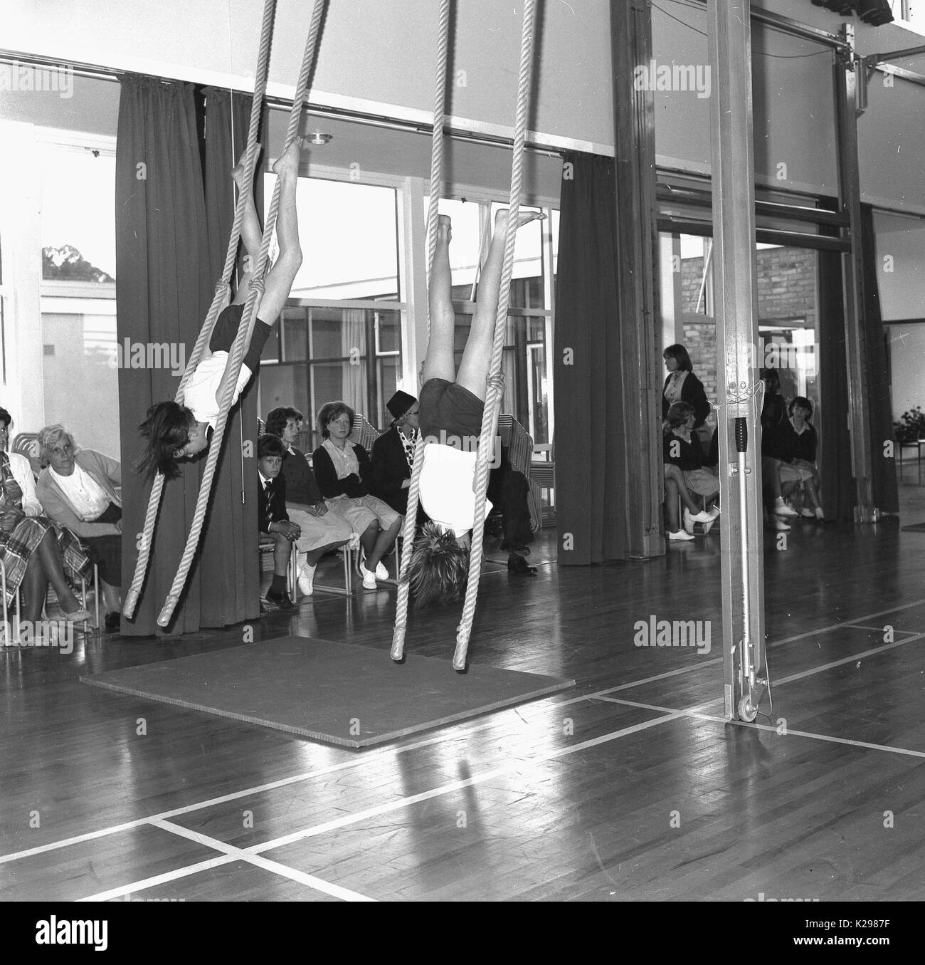 Années 1960, historique, photo montre deux gymnastes dans tradittional fille de l'école d'une trousse PE routine artistique sur les cordes d'escalade en face de parents, England, UK. Banque D'Images