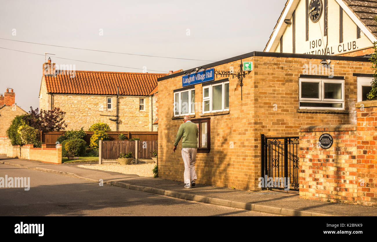 Vieux / senior man walking : monter et découper un homme âgé en prenant une marche rapide, marche quotidienne, dans des tons chauds, le soleil tôt le matin, passé un village hall en Angleterre, au Royaume-Uni. Banque D'Images