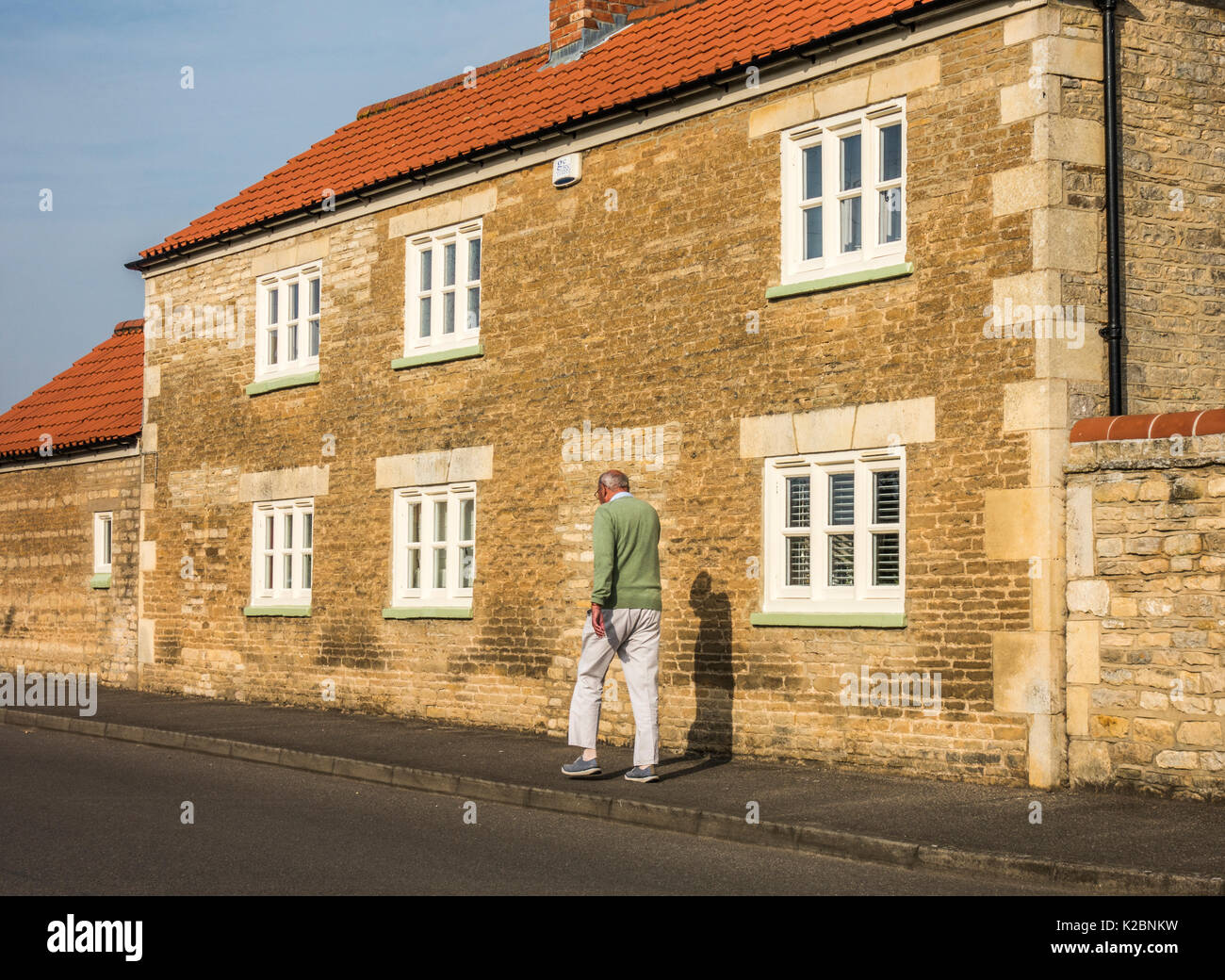 Vieux / senior man walking : monter et découper un homme âgé qui marche rapide, marche quotidienne dans le chaud soleil, tôt le matin, passé une vieille maison en pierre. Angleterre, Royaume-Uni. Banque D'Images