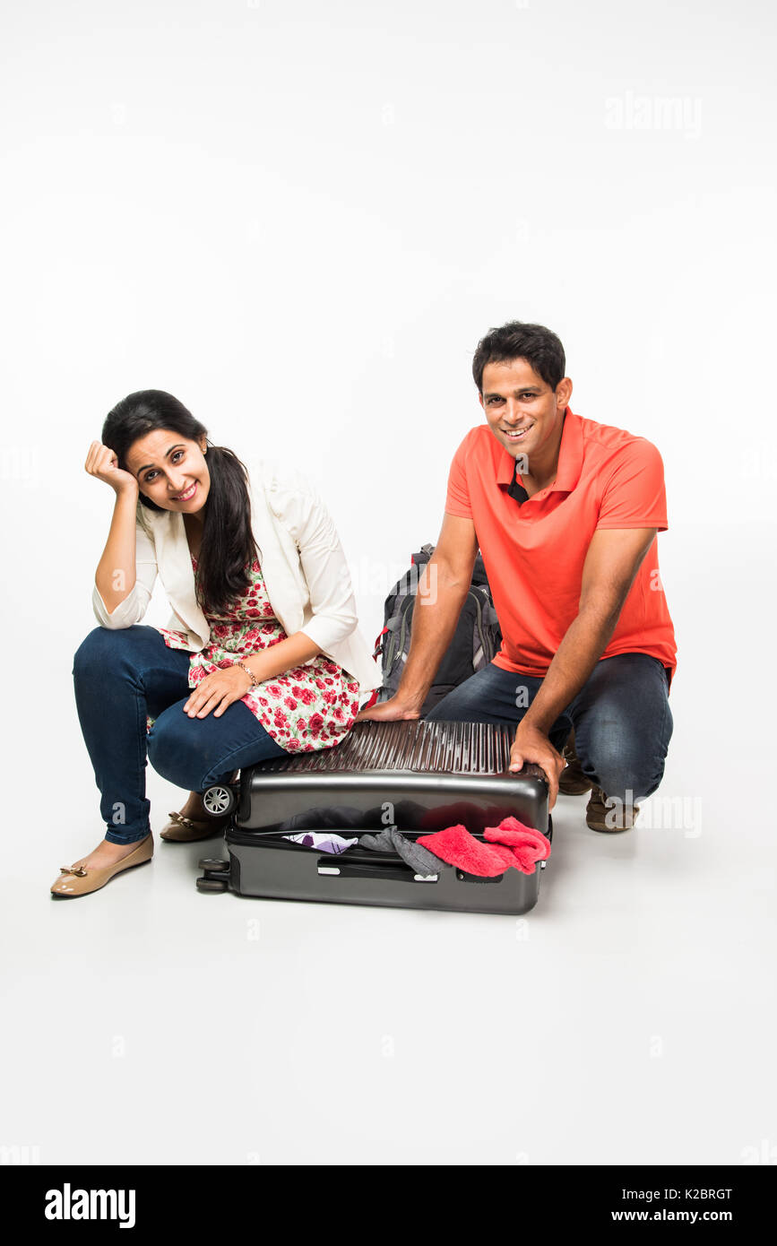 Stock photo de jeunes indiens couple packing for holiday, essayant de fermer valise pleine de vêtements, isolé sur fond blanc assis Banque D'Images