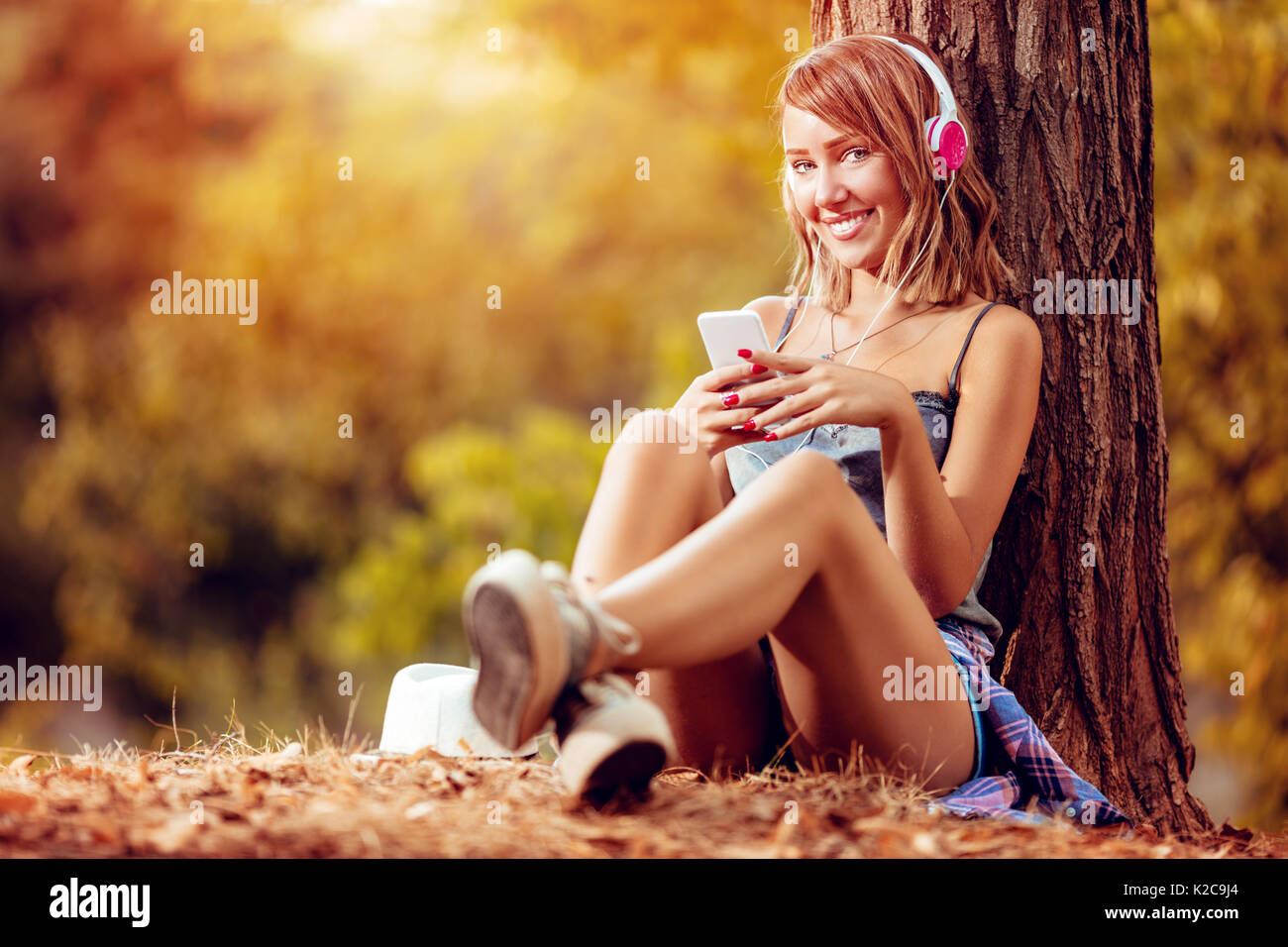 Beautiful smiling girl sitting par l'arbre dans le parc à l'automne et à l'écoute de la musique. Banque D'Images