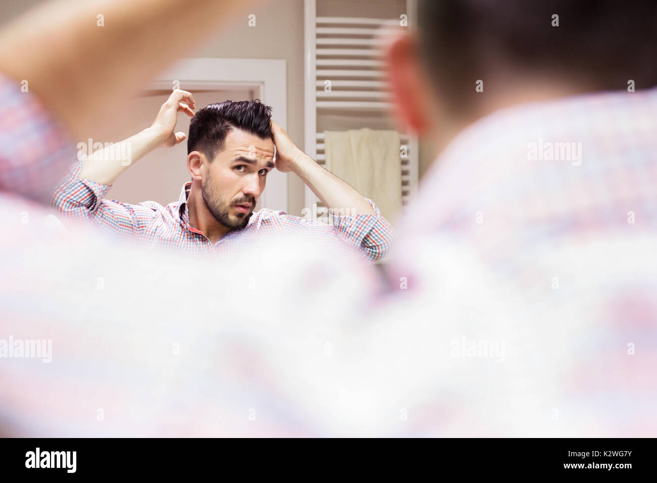 Bel homme en chemise à carreaux avec manches retroussées faisant son style de cheveux à la lumière salle de bain. Il est en se regardant dans le miroir. Banque D'Images