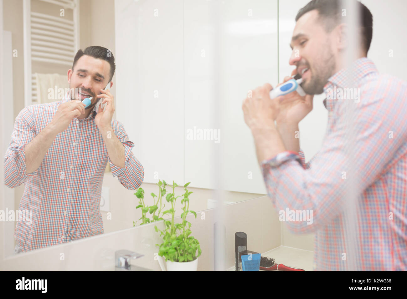 Attarctive l'homme ayant une conversation téléphonique pendant le brossage des dents. Agréable salle de bains est bien éclairée. L'homme est visible dans le miroir de la réflexion. Banque D'Images