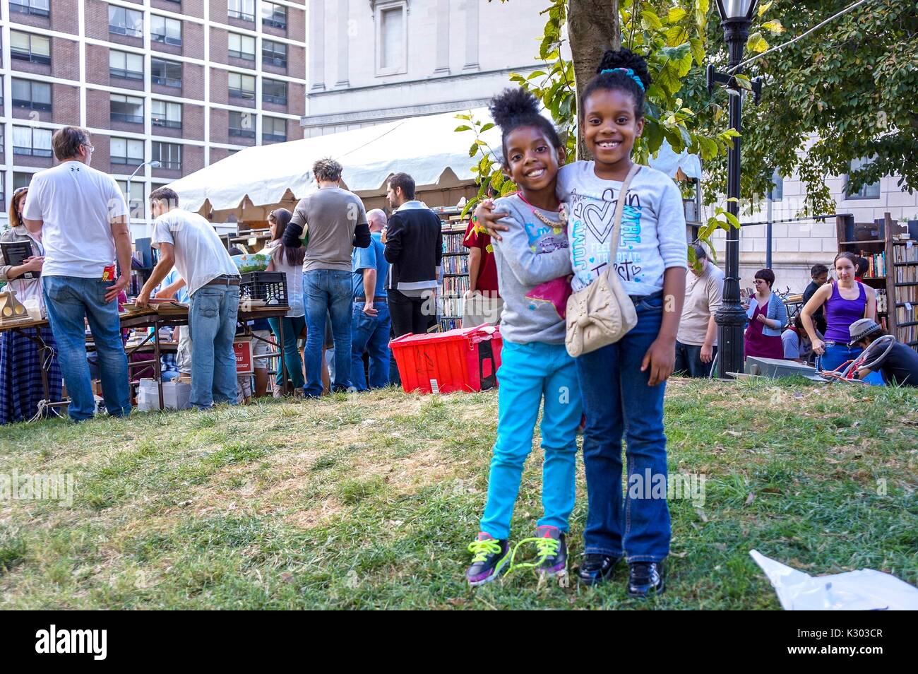 Deux jeunes sœurs se tenir avec les bras autour de l'autre tandis qu'ils posent pour leur photo, avec un arbre et plusieurs tables et de tentes de livres d'occasion à vendre durant le Festival du livre de Baltimore, Baltimore, Maryland, Septembre, 2013. Avec la permission de Eric Chen. Banque D'Images