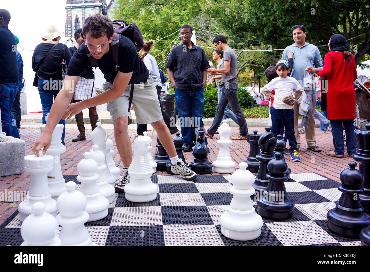 Un homme portant un sac à dos atteint pour déplacer la pièce King à partir d'un ensemble d'échecs grandeur nature, alors que les badauds derrière lui observer sa stratégie, pendant le Festival du livre de Baltimore, Baltimore, Maryland, Septembre, 2013. Avec la permission de Eric Chen. Banque D'Images