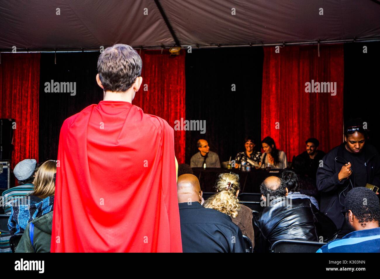 Un jeune homme avec une cape rouge est à la recherche à une discussion de groupe en face de draperies rouges avec une foule de gens assis et regarder, au Baltimore Book Festival, Baltimore, Maryland, Septembre, 2013. Banque D'Images