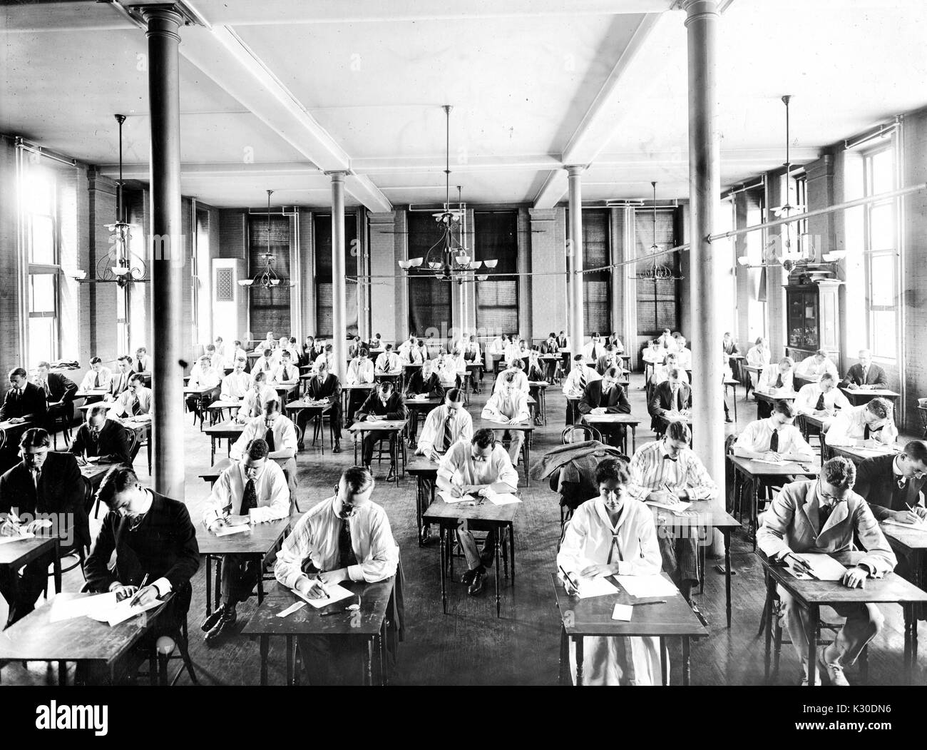 Photographie d'élèves candides portant des costumes assis à un bureau en tenant un examen dans une chambre ouverte avec de grandes fenêtres et des lustres à la Johns Hopkins University, Baltimore, Maryland, 1950. Banque D'Images
