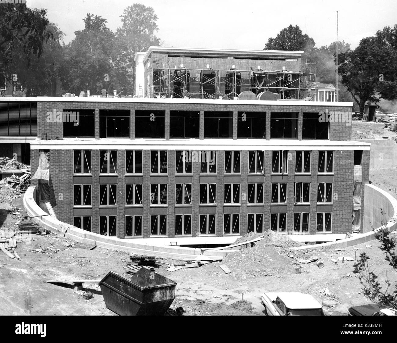 L'extérieur de la Milton S Eisenhower Library à l'Université Johns Hopkins, en construction avant son ouverture d'origine, y compris les niveaux qui s'étendent au-dessous du niveau du sol avec plusieurs fenêtres, l'intérieur d'un mur incurvé creusé dans le sol, avec une benne, échafaudage, les véhicules et les débris éparpillés autour, et Homewood House, un manoir de style fédéral du dix-neuvième siècle, visible à l'horizon, Baltimore, Maryland, août, 1964. Banque D'Images