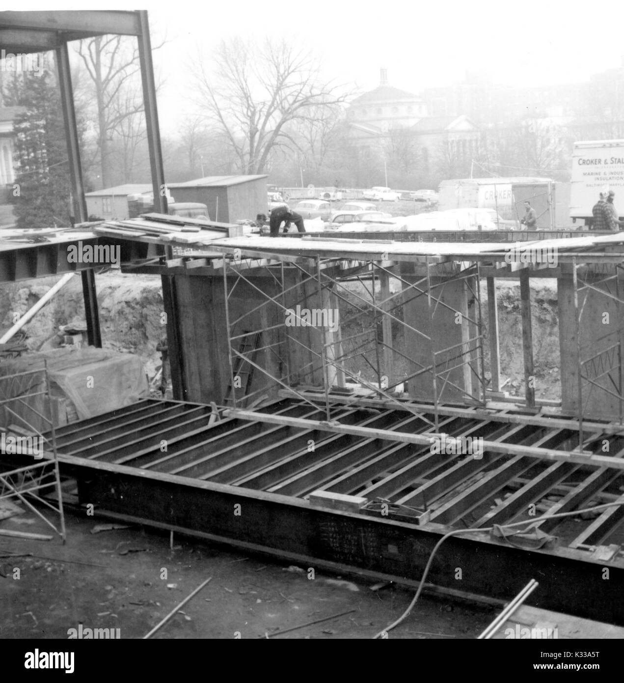 Au cours des premières étapes de la construction de la Milton S Eisenhower Library à l'Université Johns Hopkins, de grandes poutres en acier sont empilés et disposés de façon à créer la structure de Q et M niveaux, avec les travailleurs debout sur des échafaudages et de grandes planches en bois, plusieurs voitures et camions stationnés sur le terrain herbeux en face, et la rue Charles visibles à l'horizon à travers le brouillard, Baltimore, Maryland, 1963. Banque D'Images