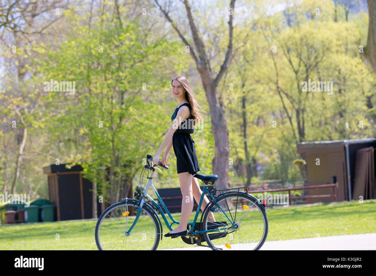 Vue Portrait of Girl sur location portant sur noir robe courte. Jeune femme heureuse équitation le long de la route sur vert printemps parc extérieur. Jeune fille sportive ri Banque D'Images