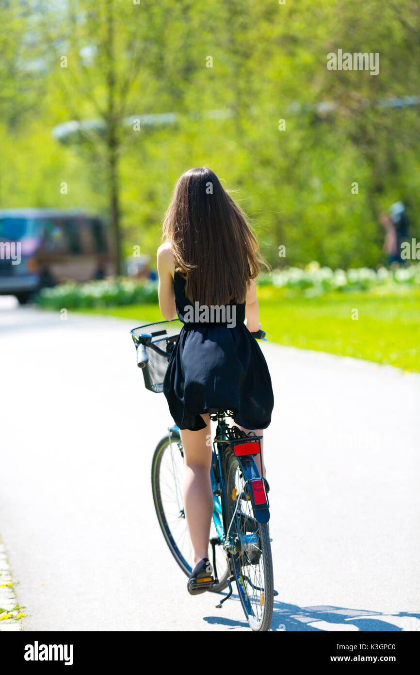 Vue arrière de la fille sur location portant sur noir robe courte. Jeune femme équitation le long de la route sur vert printemps parc extérieur. Jeune fille sportive équitation un bic Banque D'Images