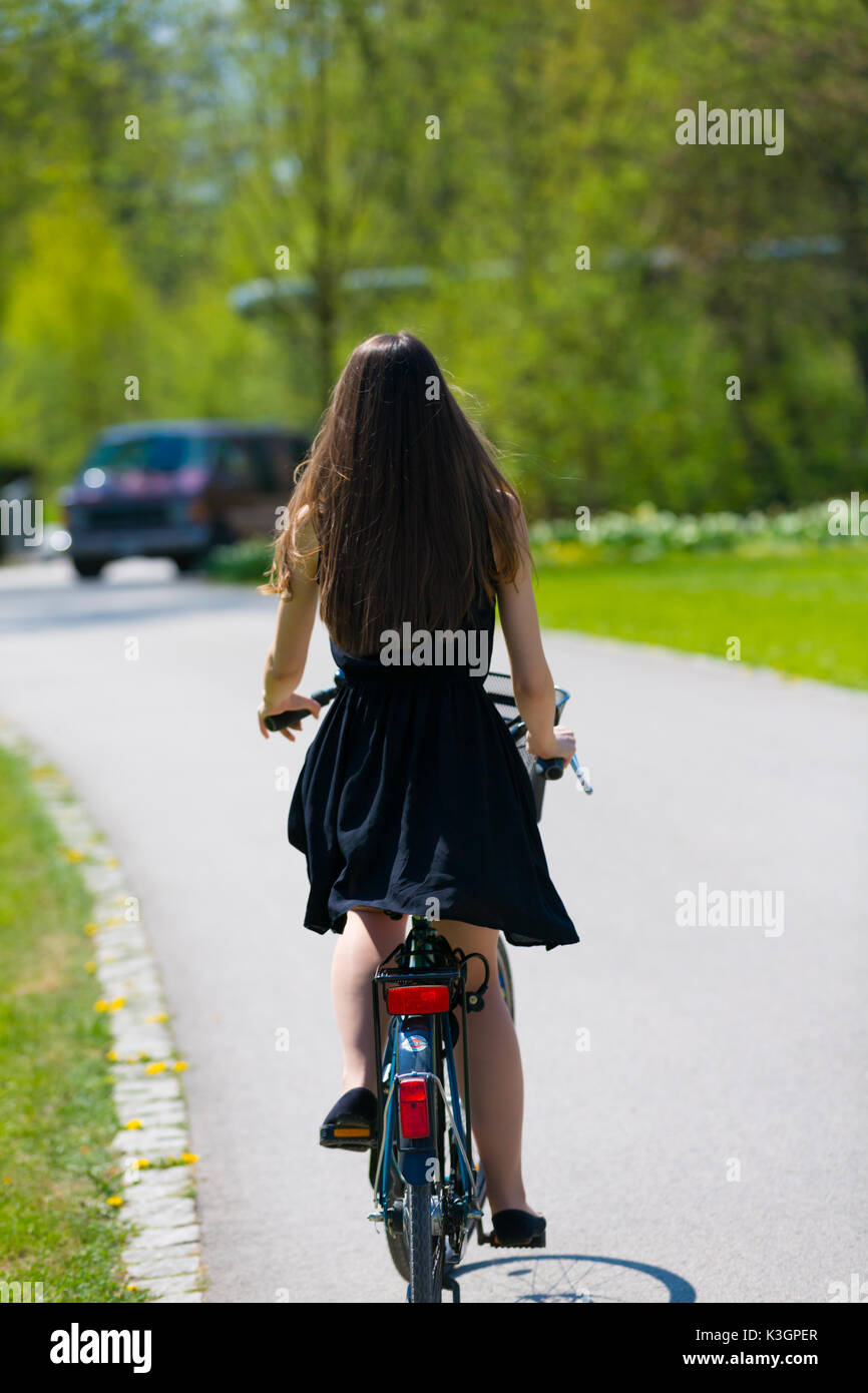 Vue arrière de la fille sur location portant sur noir robe courte. Jeune femme équitation le long de la route sur vert printemps parc extérieur. Jeune fille sportive équitation un bic Banque D'Images