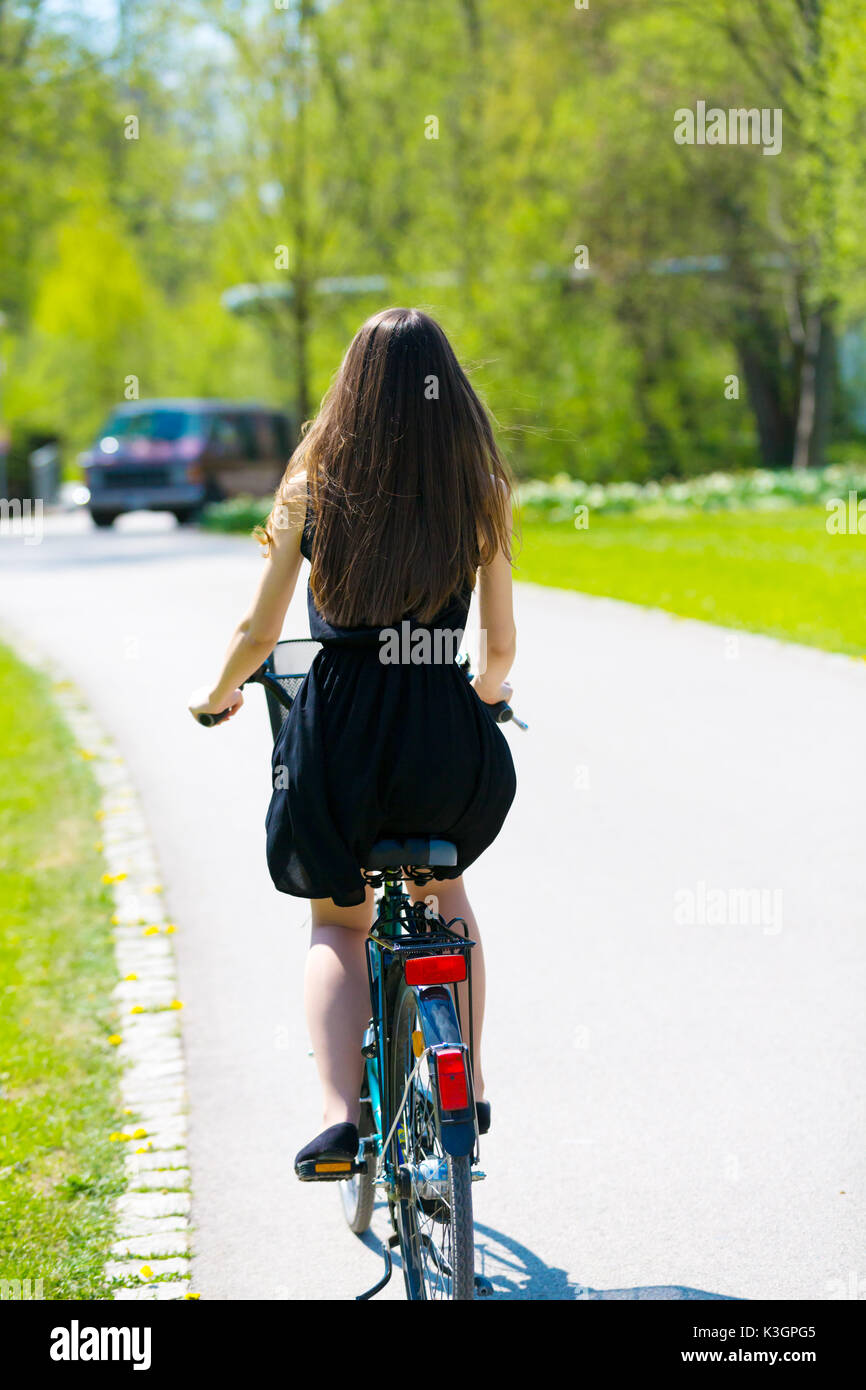 Vue arrière de la fille sur location portant sur noir robe courte. Jeune femme équitation le long de la route sur vert printemps parc extérieur. Jeune fille sportive équitation un bic Banque D'Images