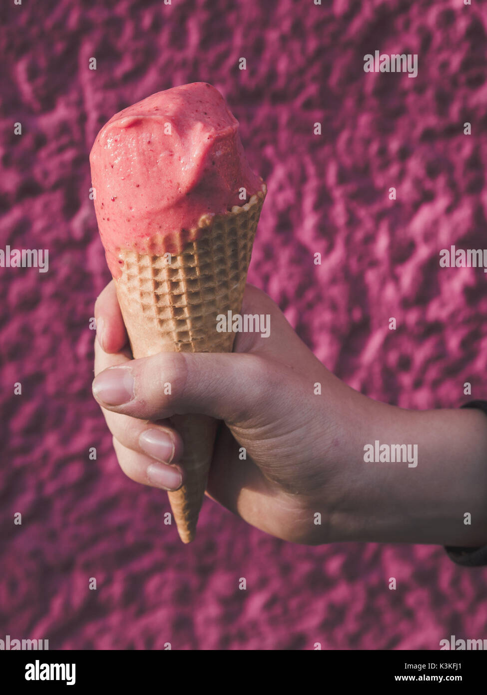 Main d'un garçon tenant un cornet de crème glacée aux fraises sur fond rose Banque D'Images