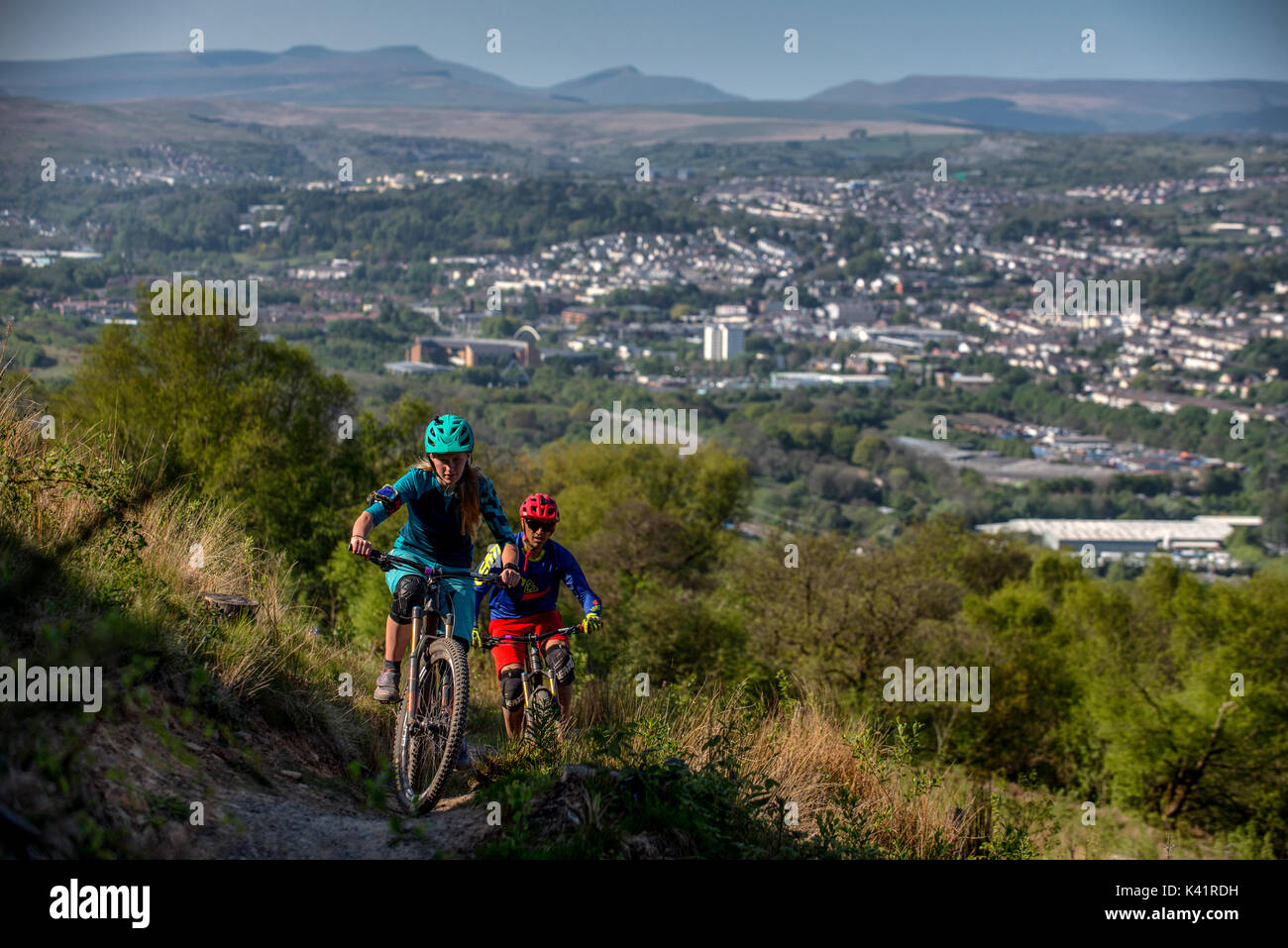 Un vélo de montagne femmes mène un homme une montée au Bikepark de galles avec la ville de Merthyr Tydfil et les Brecon Beacons dans l'arrière-plan. Banque D'Images