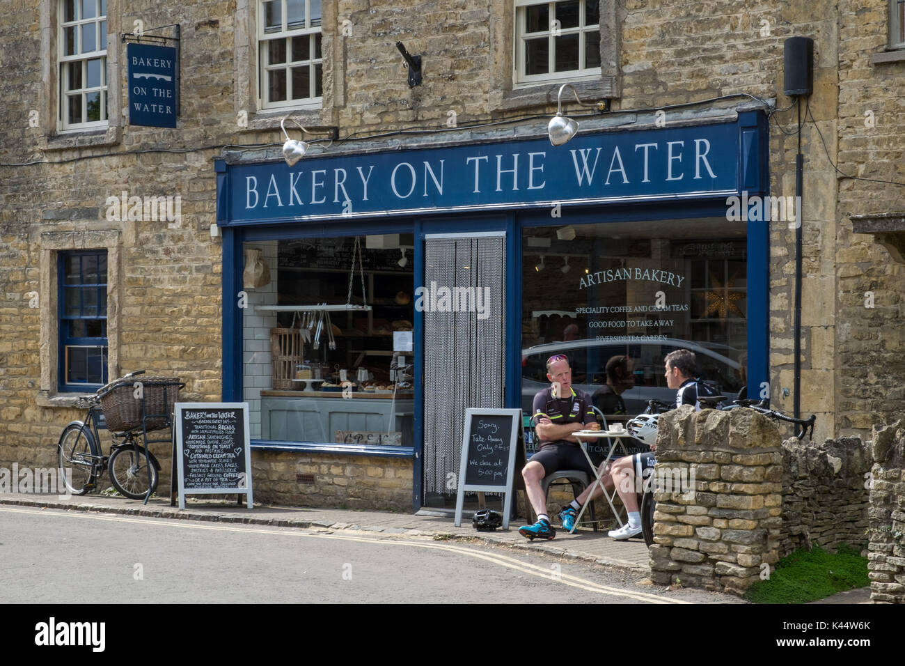 Les touristes à vélo profiter du soleil en été, un café et une collation à l'extérieur d'une boulangerie artisanale dans l'historique village de Cotswold bourton on the water Banque D'Images