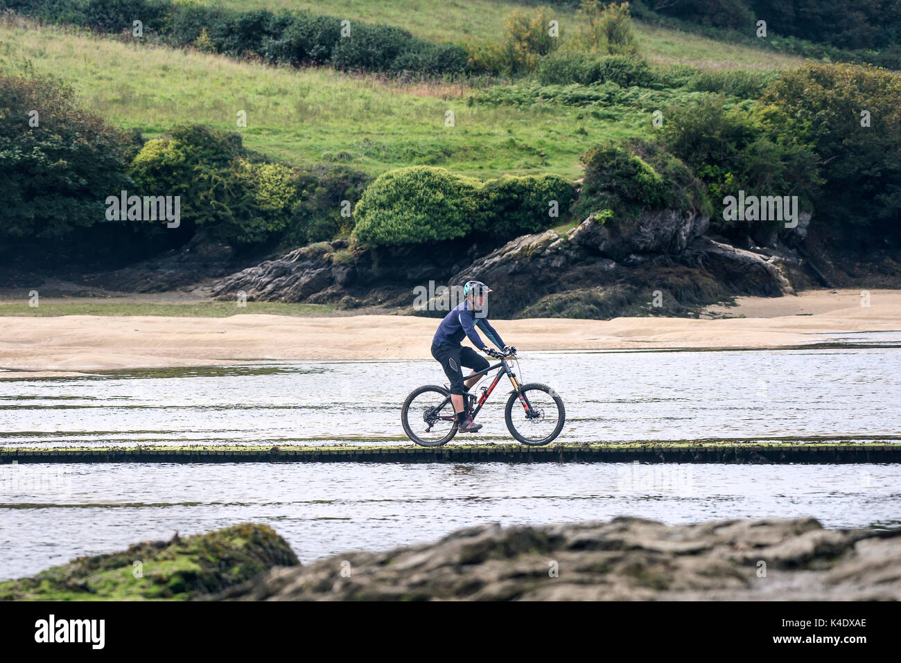 Estuaire Gannel - un homme monté sur un vélo de montagne, traversée de la rivière Gannel par une passerelle à marée basse. Banque D'Images