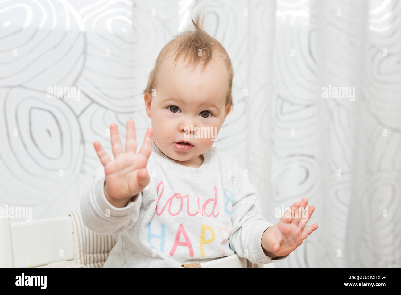 Onze mois baby girl sitting dans un tabouret pour faire des grimaces à la caméra, elle élève ses deux mains en l'air Banque D'Images