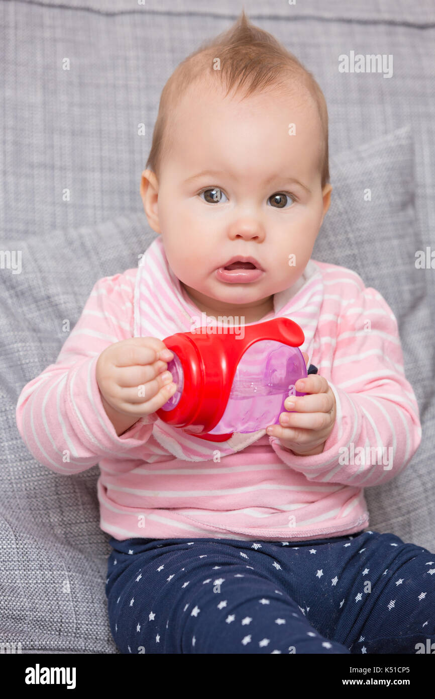 Baby Girl holding red bouteille d'eau en plastique, assis sur un canapé gris Banque D'Images