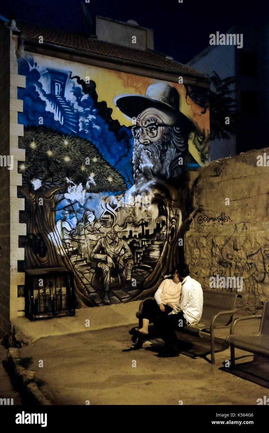 Un couple juif orthodoxe assis dans la nuit dans un banc en face d'un mur peint représentant le rabbin aryeh levin appelé aryeh du CER, qui était un rabbin orthodoxe surnommé le "père des prisonniers' pour ses visites aux membres de l'underground juifs emprisonnés à la prison centrale de Jérusalem dans le quartier russe durant le mandat britannique. situé dans l'ancien quartier cour sukat shalom à Nachlaot ou nahlaot un regroupement de 23 quartiers ancienne cour qui peuplaient la nouvelle ville après avoir quitté la ville fortifiée de Jérusalem à la fin du 19e siècle situé dans l'ouest de Jérusalem israël Banque D'Images