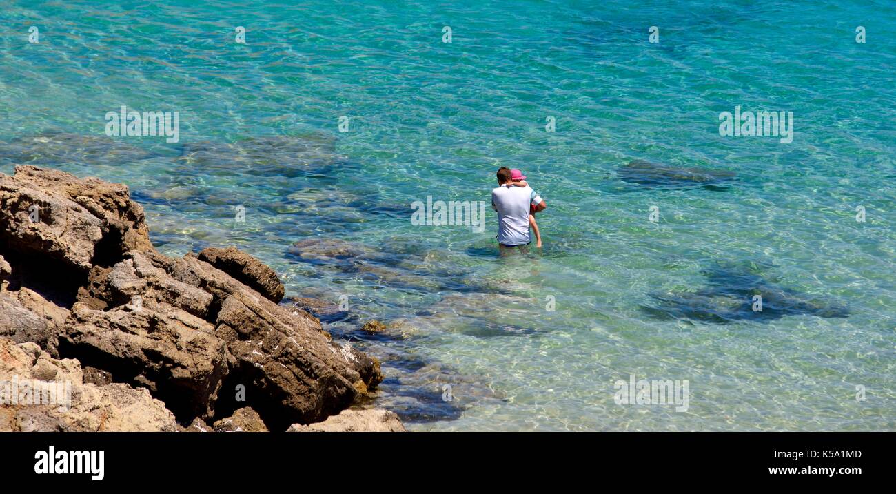 Un homme portant un enfant par l'eau de mer peu profonde Minorque Minorque espagne Banque D'Images