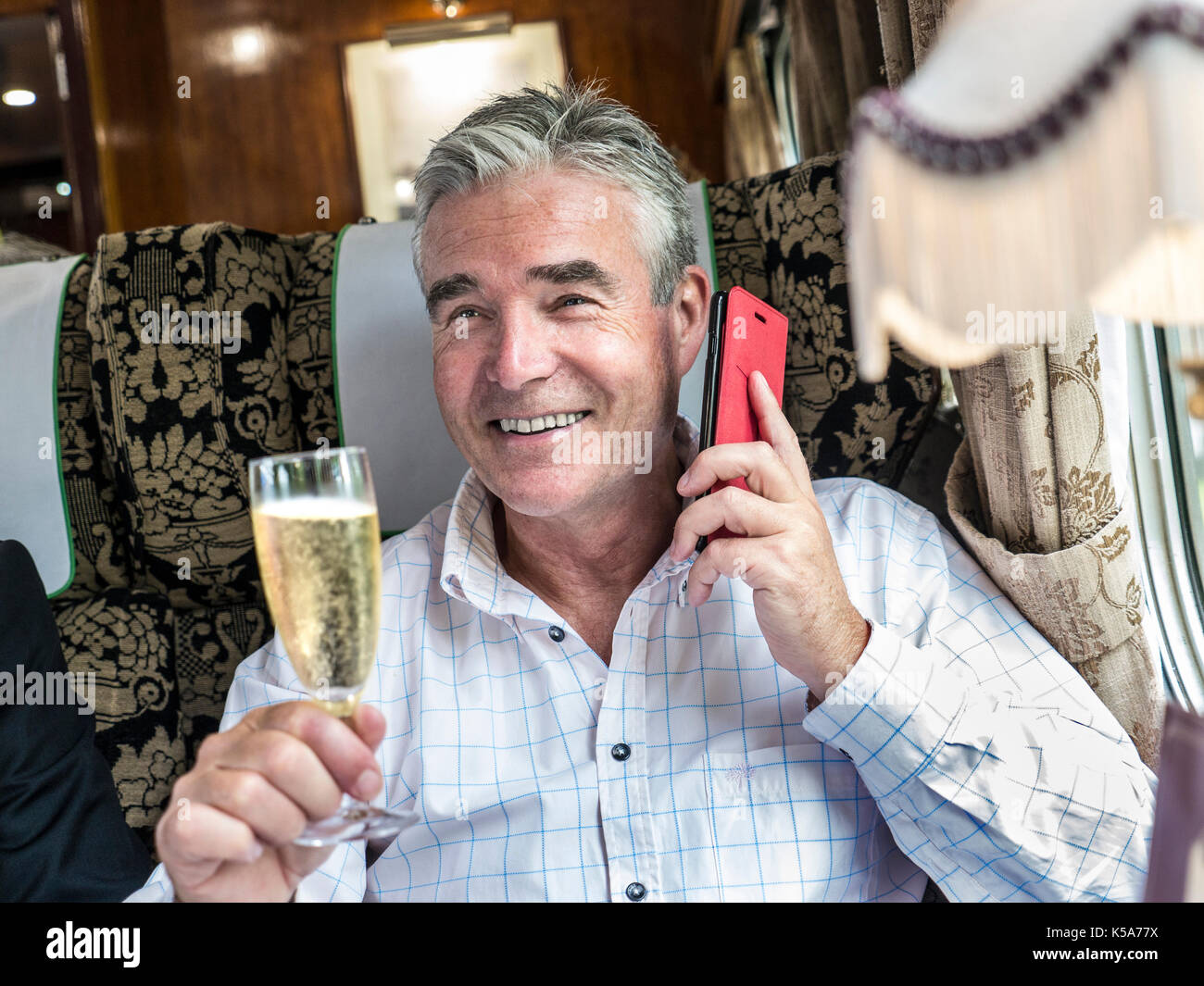 TRAIN DE LUXE DE PREMIÈRE CLASSE CHAMPAGNE décontracté heureux homme mature, parlant sur son smartphone en profitant de son voyage dans la calèche de chemin de fer de table Pullman Banque D'Images