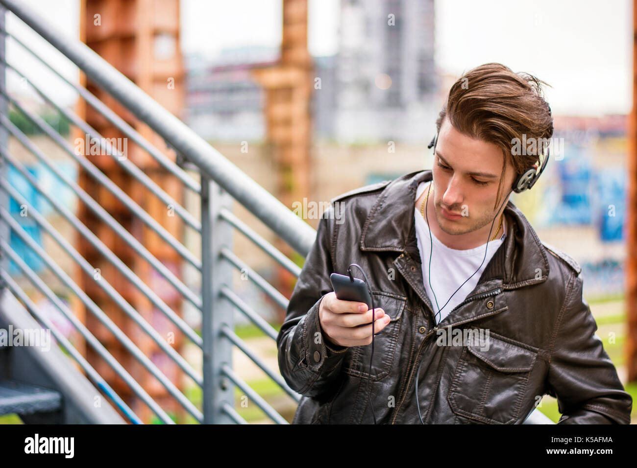 Beau jeune homme debout à l'extérieur dans l'environnement urbain sur les escaliers en métal, using cell phone Banque D'Images