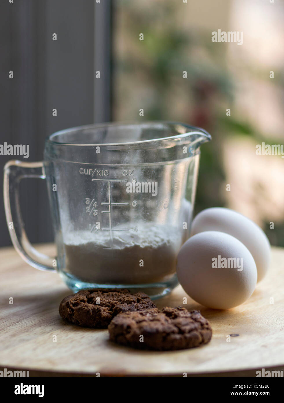 Une tasse de mesure en verre, deux œufs et chocolat cookies cuits sur le dessus d'une planche en bois, avec une lumière douce et non mise au point historique Banque D'Images