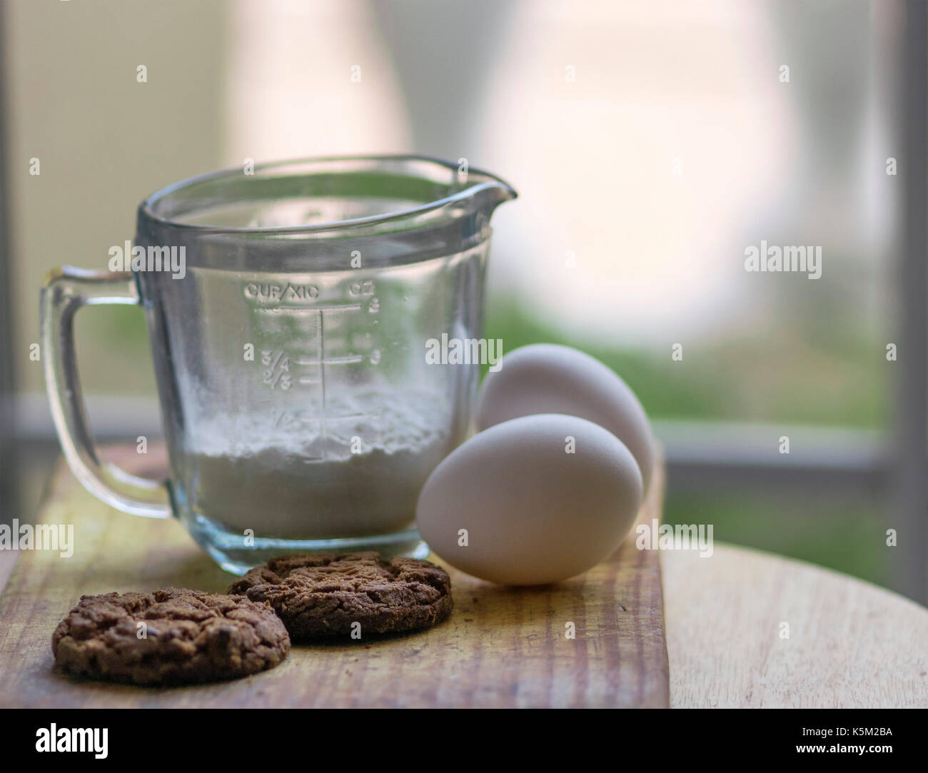 Une tasse de mesure en verre, deux œufs et chocolat cookies cuits sur le dessus d'une planche en bois, avec une lumière douce et non mise au point historique Banque D'Images
