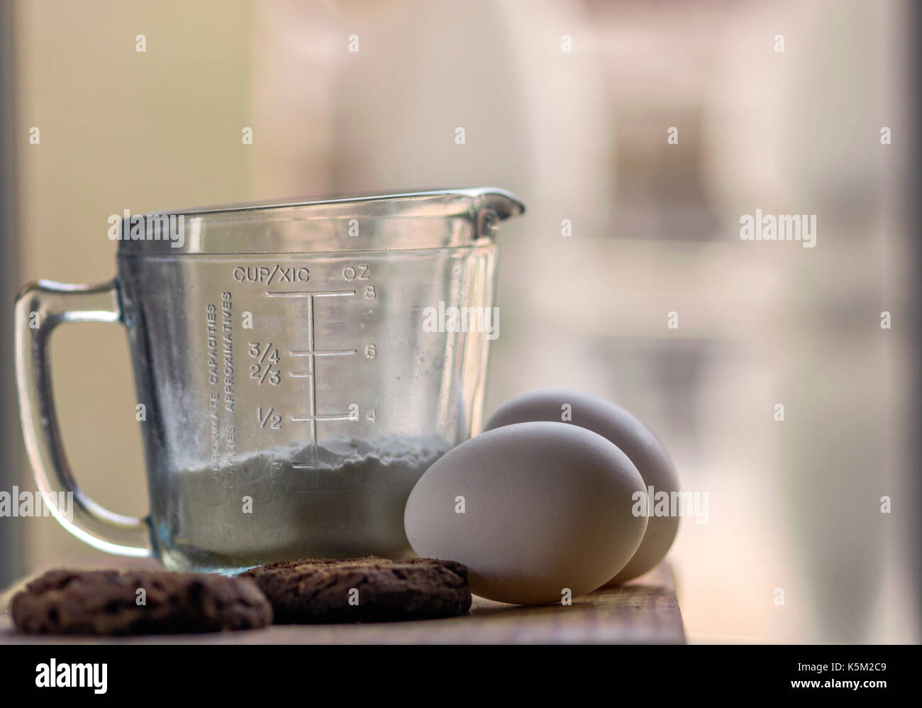 Une tasse de mesure en verre, deux œufs et chocolat cookies cuits sur le dessus d'une planche en bois, avec une lumière douce et non mise au point historique Banque D'Images