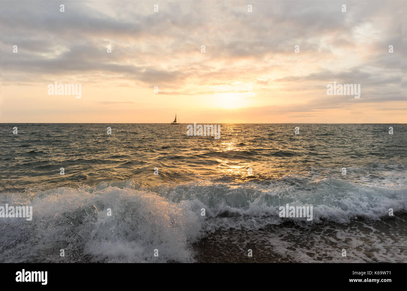 Voilier coucher du soleil est un voilier naviguant le long de l'océan au coucher du soleil avec une vague s'écraser sur le rivage. Banque D'Images