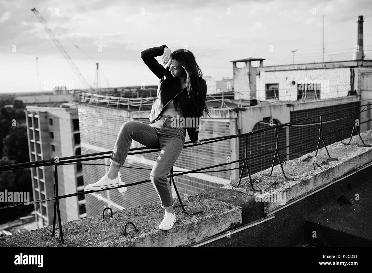 Portrait d'une magnifique jeune femme en noir veste en cuir, jeans et baskets assis sur les mains courantes sur le toit avec vue pittoresque d'un parc. noir Banque D'Images