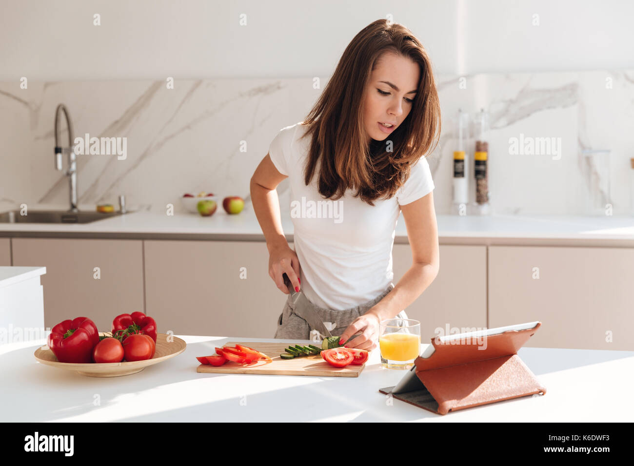 Jeune femme concentré couper des légumes sur une planche en bois et à la tablette à l'ordinateur pendant la cuisson dans une cuisine Banque D'Images