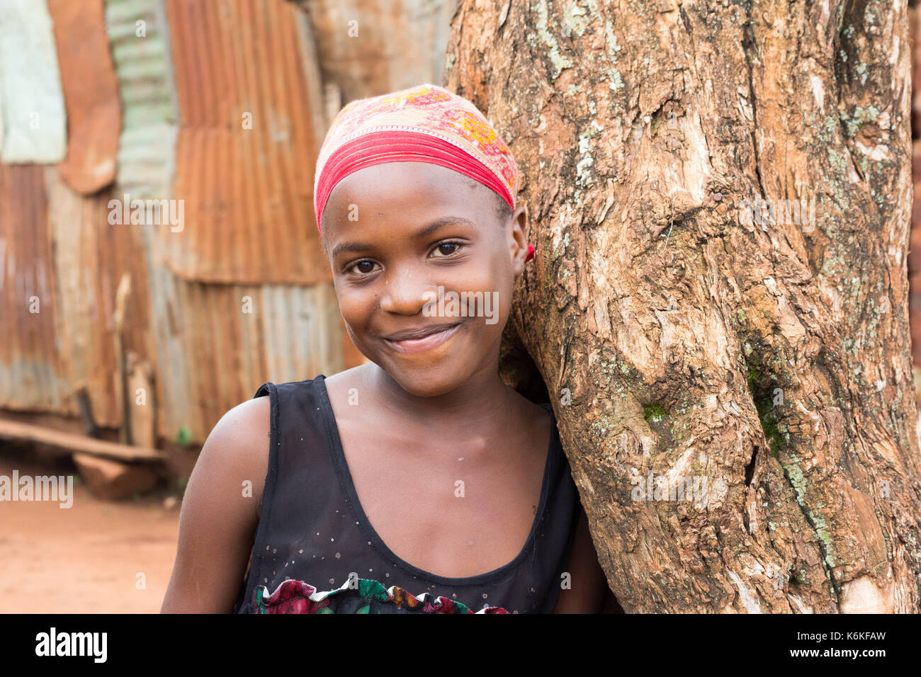 Une belle black teenage girl smiling et appuyé contre un arbre. elle porte un foulard rouge. Banque D'Images
