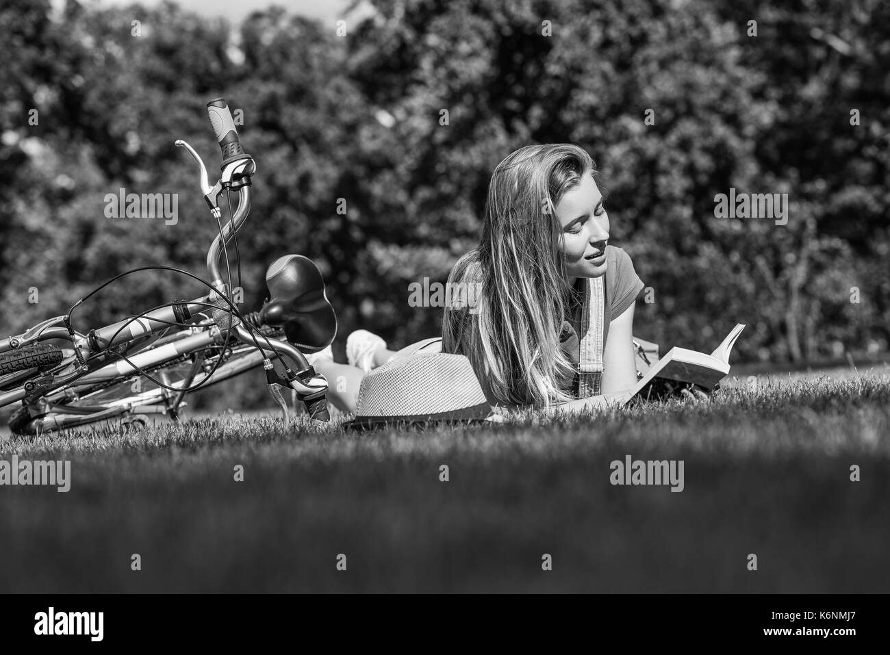Shot monochrome d'une jeune belle femme appréciant la lecture d'un livre allongé dans l'herbe près de son vélo se reposer au soleil sur une chaude journée d'été nature Banque D'Images