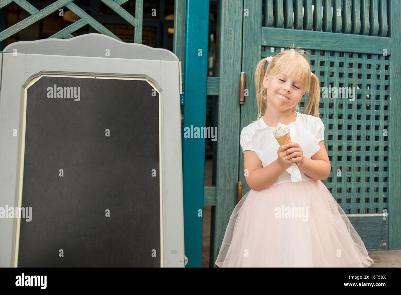 Peu cute blonde girl dans une belle robe de manger des glaces à l'extérieur. Il y a un tableau noir avec place pour le texte sur une photo. Banque D'Images