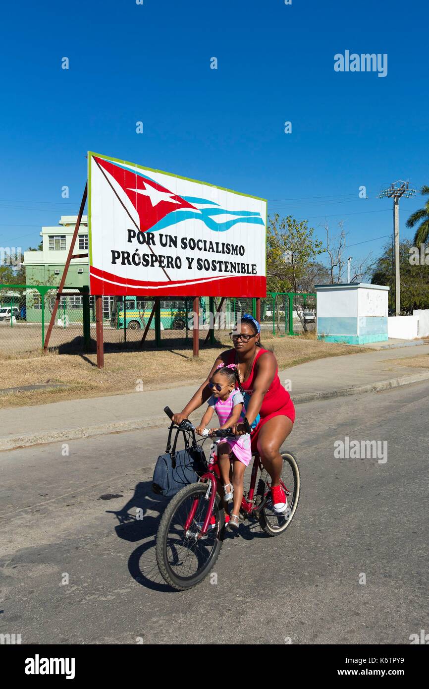 Cuba, Cienfuegos, Cienfuegos province, Punta Gorda, femme et enfant sur un vélo à l'avant d'un signe de la propagande politique Banque D'Images