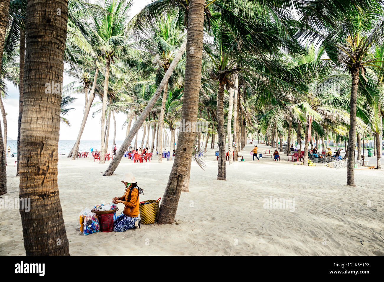 Femme avec chapeau et robe orange la préparation et la vente des aliments sur la plage de sable et de palmiers à Hoi An, Vietnam Banque D'Images