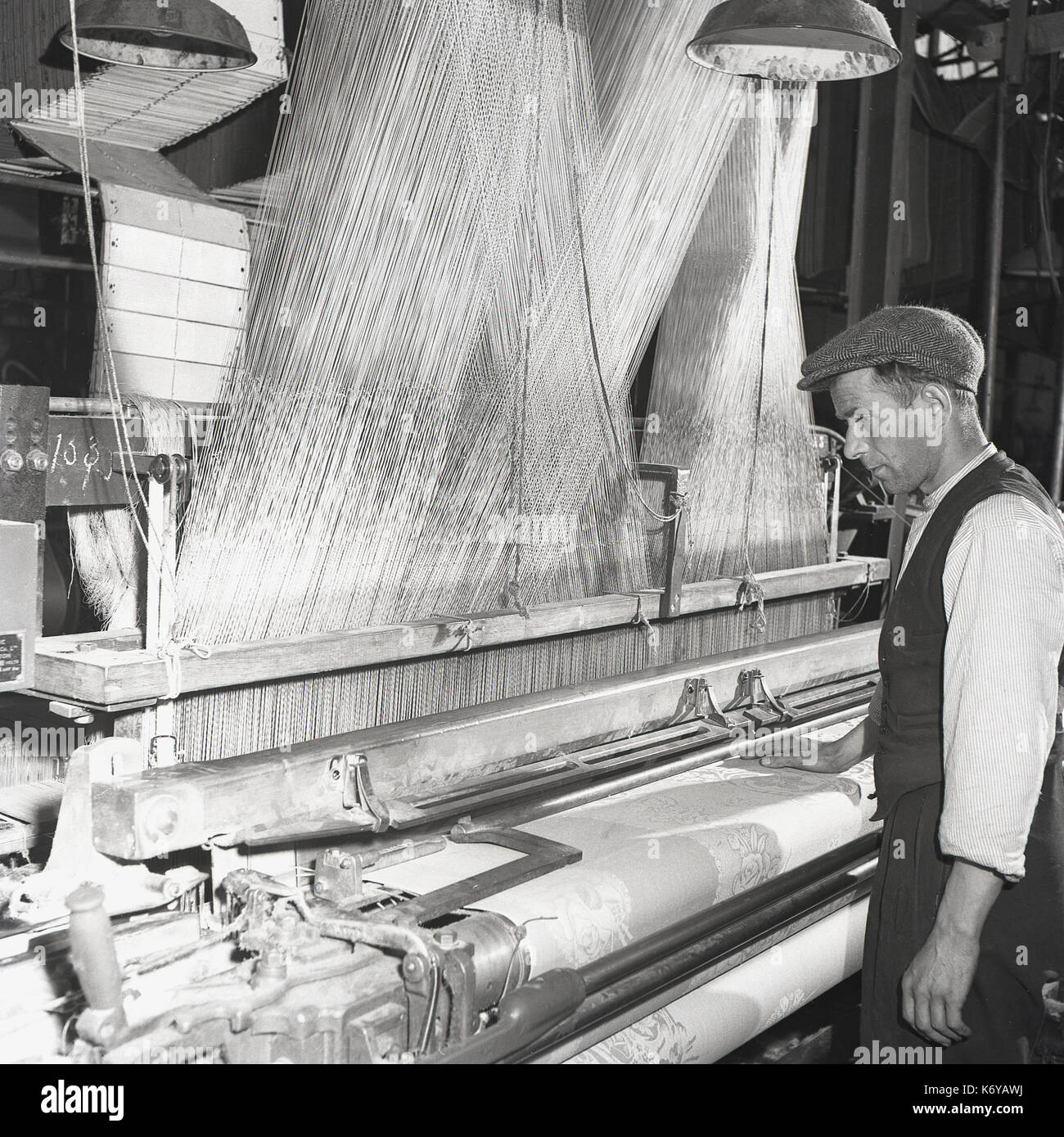 Années 1950, l'Irlande du Nord, un homme travailleur du textile dans un gilet et cap à l'aide d'un métier mécanisé ou cadre pour faire le tissu en lin de la laine, du lin threads. Banque D'Images